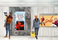 Digital Signage Display Promoting Black Friday Sales: A vibrant digital signage screen showcasing a bold "Black Friday Sale" message with bright colors, dynamic graphics, and a list of highlighted discounts. The display includes a QR code for exclusive offers and is positioned in a busy retail environment with shoppers in the background.